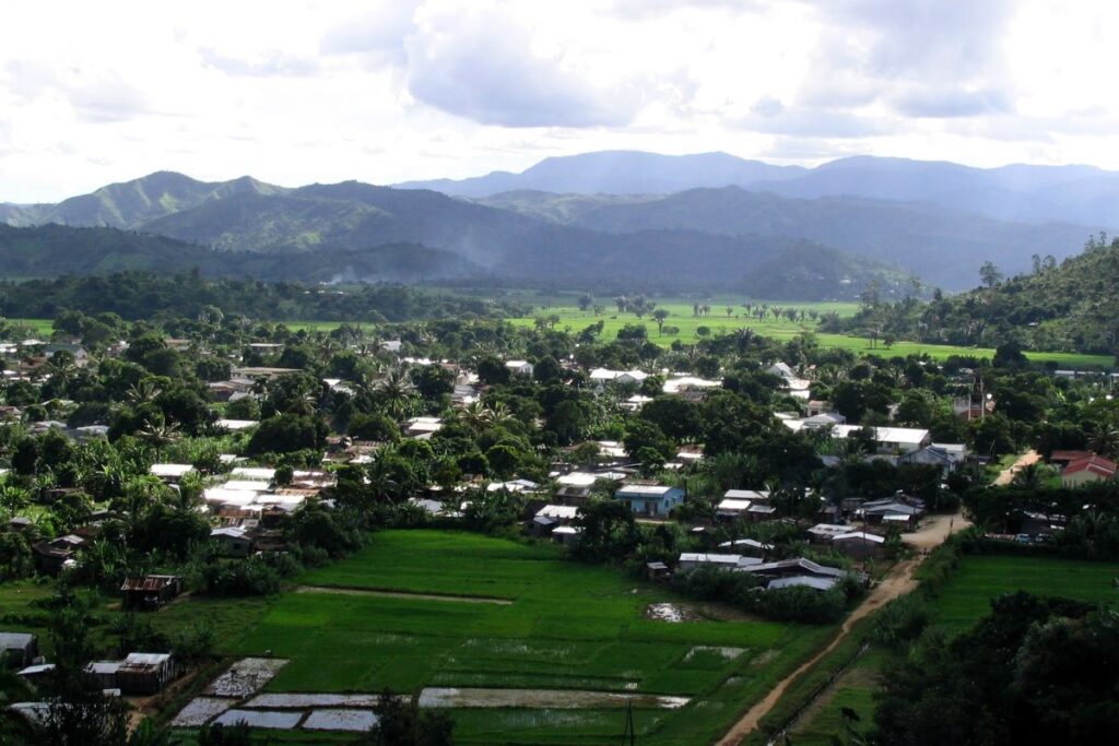 Trano kely vita amin'ny vy no mivondrona eo anivon'ny hazo, miaraka amin'ny tanimbary maitso any ankoatra ary tendrombohitra rakotra ala miakatra eny lavitra eny.